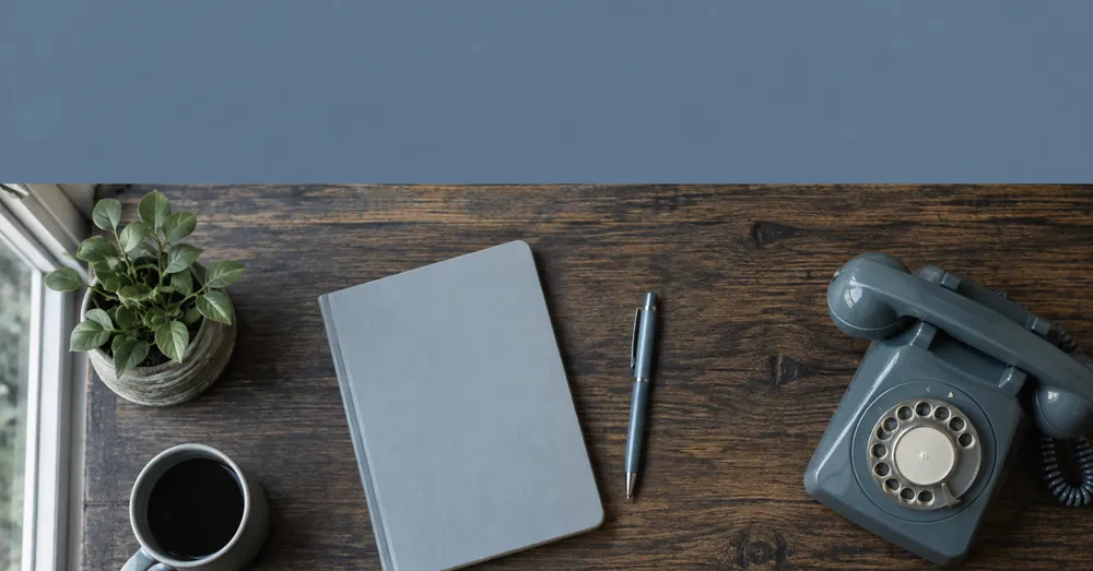Mesa pequena e organizada vista de cima, com caderno, caneta e telefone de mesa.